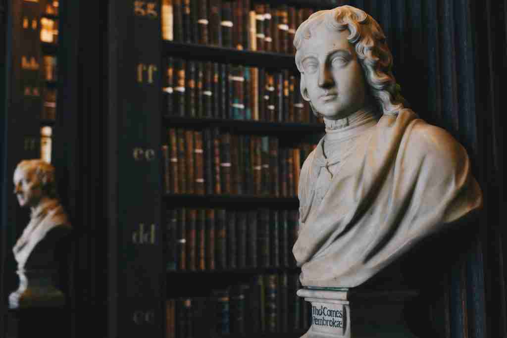 Bustos de mármol sobre pedestales en una biblioteca poco iluminada con hileras de libros antiguos al fondo, evocando una atmósfera que recuerda a los Hijos de Dios de la Biblia.