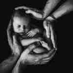 Imagem em preto e branco de um bebê recém-nascido embalado por quatro mãos contra um fundo escuro, lembrando o apoio e a união encontrados entre os filhos de Leah na Bíblia.