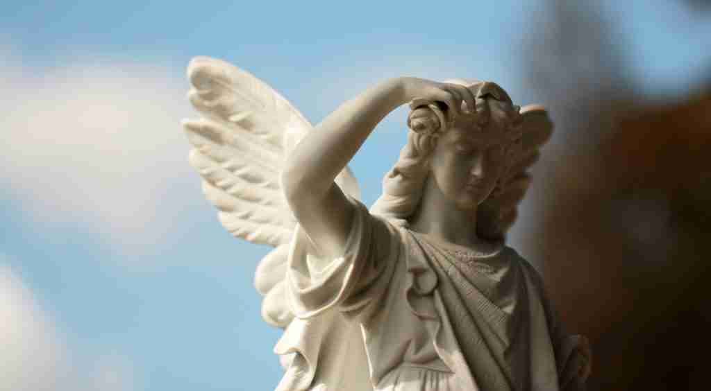 Un primer plano de un ángel de piedra blanca con alas y expresión serena, sobre un cielo azul con ligeras nubes, evoca a los Hijos de Dios de la Biblia.