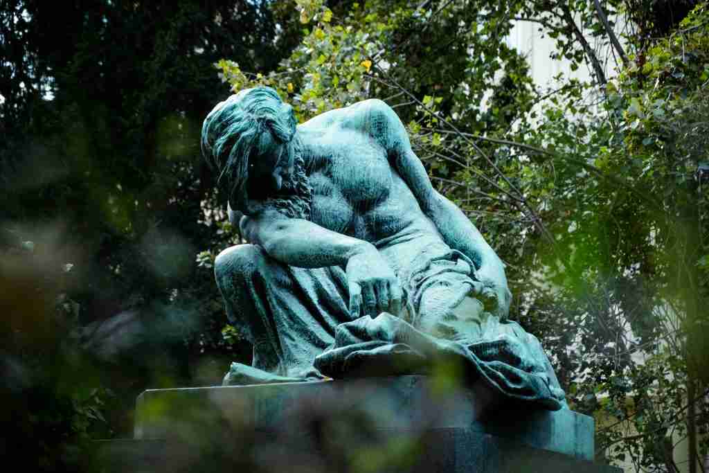 Una estatua sombría y desgastada de una figura sentada con la cabeza inclinada, rodeada de exuberante follaje en un entorno tranquilo al aire libre, evoca la reverencia que se encuentra en las historias de los Hijos de Dios en la Biblia.