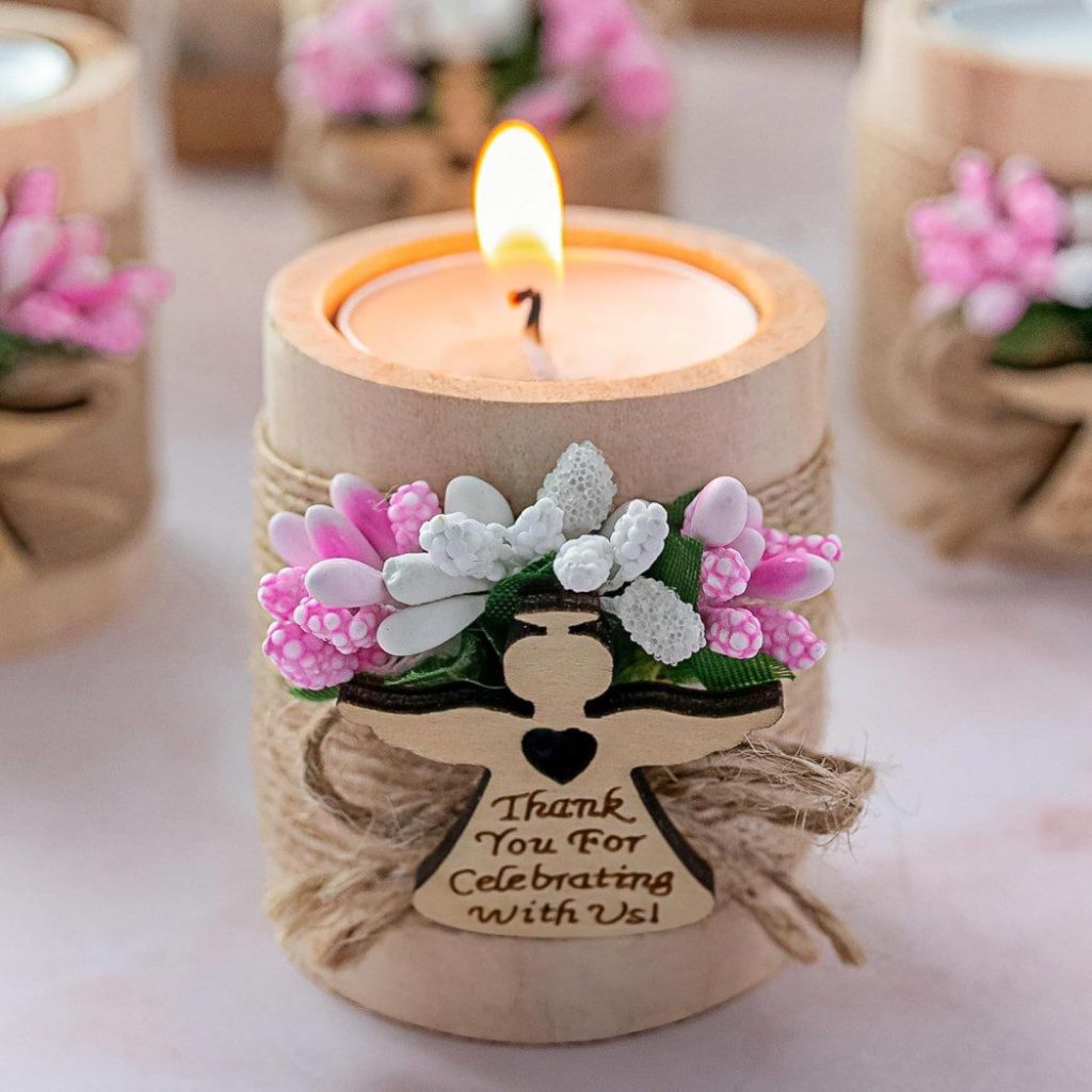 Esta vela decorativa, adornada con flores rosas y blancas, cordel y una cruz de madera que dice "Gracias por celebrarlo con nosotros", es una idea perfecta para regalar en un bautizo.