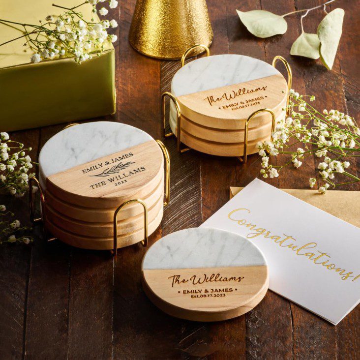 Posavasos de mármol y madera con grabados personalizados, rodeados de flores, una caja de regalo y una tarjeta de felicitación son ideas para regalar en un bautizo.