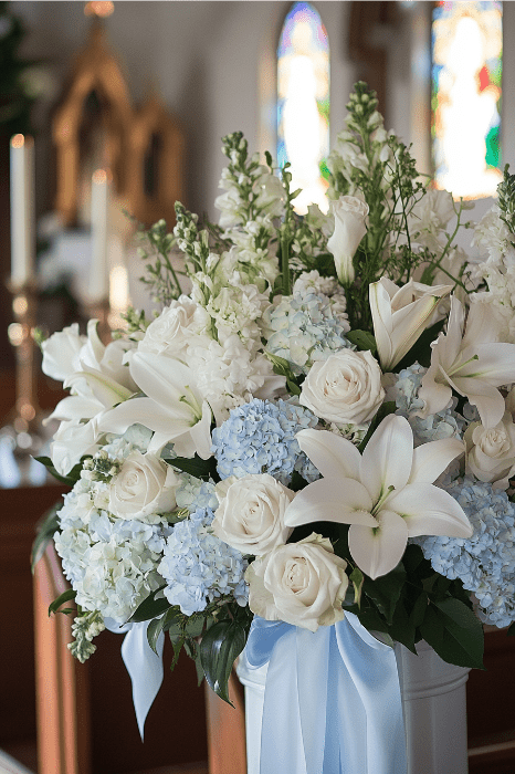Elegantes ideas de arreglos florales para altares de iglesias cobran vida con un ramo de lirios blancos, rosas y hortensias azules. Delicadas cintas se mezclan a la perfección en esta impresionante exhibición, con un telón de fondo de vidrieras para un ambiente sereno y cautivador.