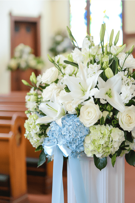 Un impresionante arreglo floral para altares de iglesias, este ramo incluye lirios blancos, rosas y hortensias azules, todo elegantemente atado con una cinta azul para complementar el sereno entorno de la iglesia.