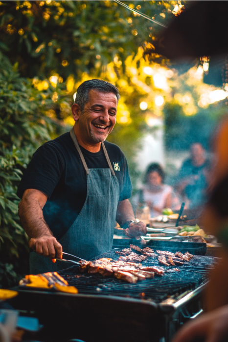Um homem sorrindo enquanto cozinha carne em uma churrasqueira ao ar livre, cercado por amigos comemorando o Dia dos Pais sob a sombra das árvores, enquanto poemas sinceros são trocados entre eles, acrescentando um toque de calor e espiritualidade à reunião.