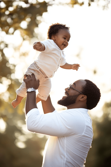 Um homem sorridente de branco levanta um bebê feliz ao ar livre, lembrando uma cena de poemas sinceros do Dia dos Pais, com árvores abraçando-os graciosamente ao fundo.