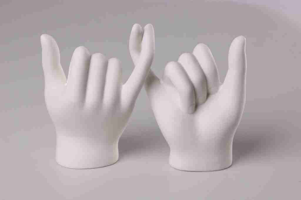 ¿Qué son las Promesas de AA? (Libro Grande de Alcohólicos Anónimos) Dos esculturas de manos de cerámica blanca, una con el signo de la paz y la otra con el pulgar hacia arriba, simbolizan las oraciones y promesas de AA sobre un fondo liso.