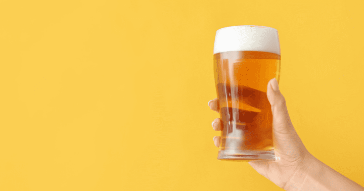 Uma mão segura uma caneca de cerveja âmbar com espuma, lembrando celebrações antigas, em um sólido pano de fundo amarelo.