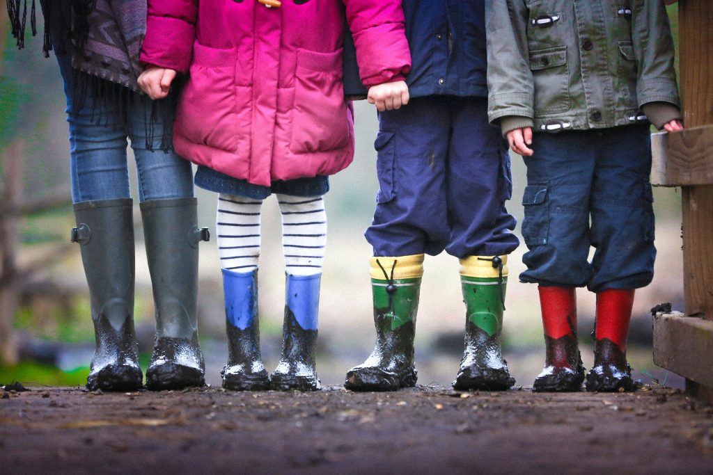 Cuatro niños, recién salidos de una alegre lección sobre la fe en la escuela dominical, se colocan en fila con coloridas botas de lluvia, mostrando orgullosos sus pies cubiertos de barro sobre la superficie mojada.
