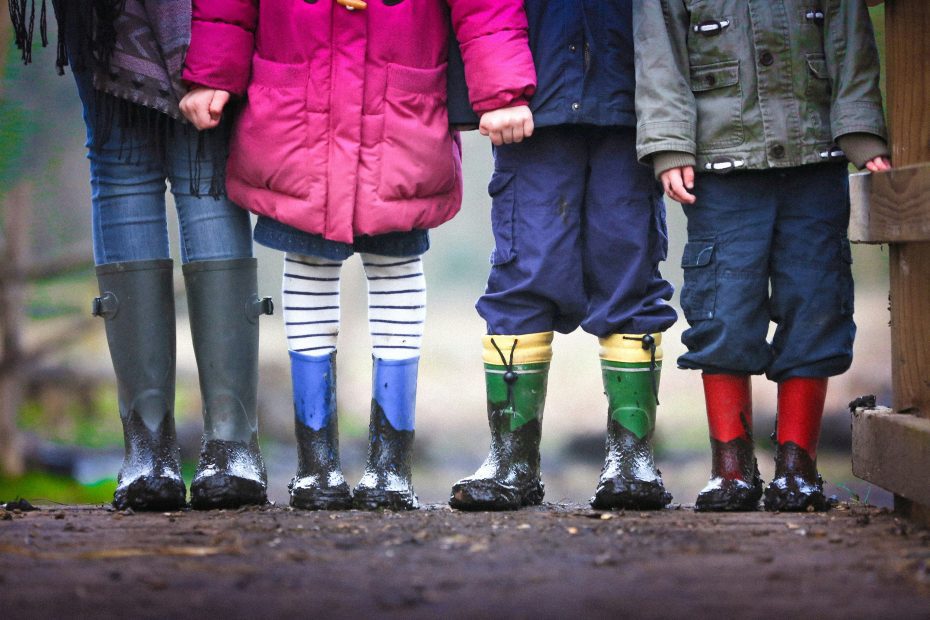 Quatro crianças, recém-saídas de uma alegre aula da Escola Dominical sobre fé, estão em uma fileira usando botas de chuva coloridas, exibindo orgulhosamente seus pés cobertos de lama na superfície molhada.