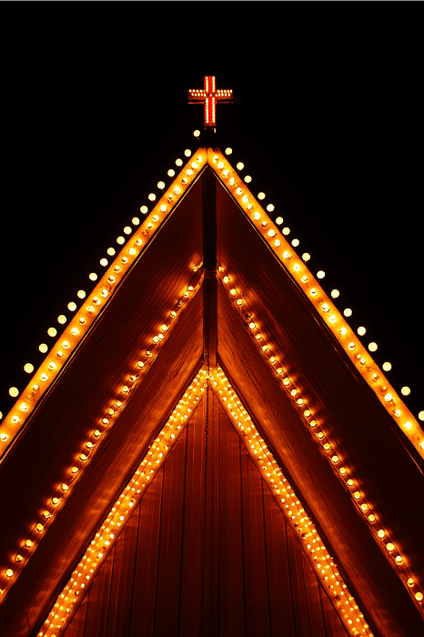 Um telhado de estrutura em A, iluminado por luzes alaranjadas e coroado por uma cruz iluminada, ergue-se orgulhosamente contra o céu escuro da noite - uma cena que lembra poemas para o aniversário de uma igreja que celebra a unidade e a fé.