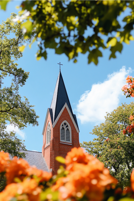 Um pitoresco campanário de igreja com uma cruz, emoldurado por árvores verdes e flores alaranjadas sob um céu azul, evoca a beleza atemporal encontrada em poemas para comemorações de aniversário de igreja.
