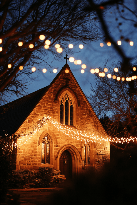 Uma igreja de pedra calorosamente iluminada ao anoitecer, adornada com luzes brilhantes, cria um cenário poético para o aniversário da igreja. Cercada por árvores nuas de inverno, ela sussurra versos silenciosos de fé e celebração no ar fresco da noite.