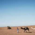 Uma pessoa vestida de azul conduz dois camelos por um vasto deserto arenoso sob um céu azul claro, lembrando as jornadas atemporais dos contos bíblicos, como quando Rebeca partiu em uma tenra idade para se casar com Isaac.