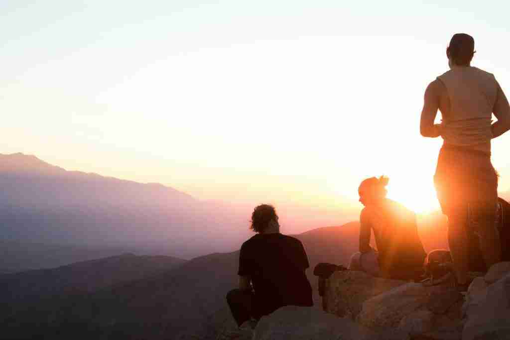 Tres personas sentadas en un saliente rocoso, reflexionando en el sereno abrazo del paisaje montañoso al atardecer, sus pensamientos entrelazados con las oraciones de los Pasos de AA.