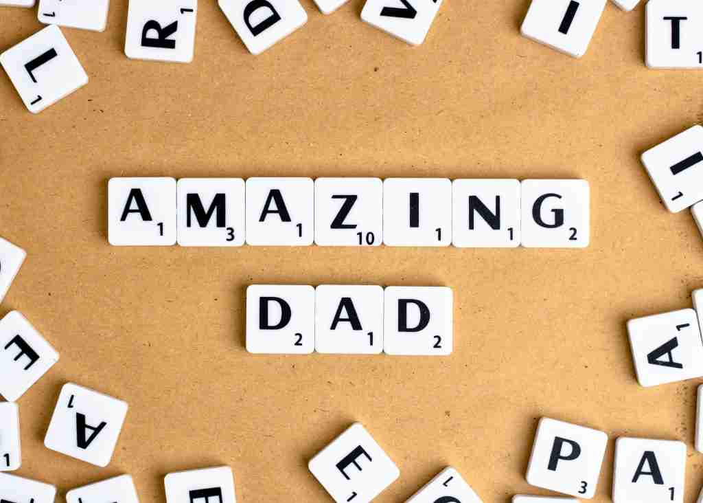 Peças de Scrabble dispostas de forma a soletrar "AMAZING DAD" em um fundo marrom, com peças espalhadas, o que o torna uma ideia perfeita para o presente do Dia dos Pais.