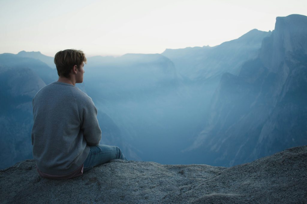 Una persona está sentada en una roca, contemplando el vasto paisaje montañoso bajo un cielo azul y brumoso, donde la grandeza de la naturaleza se siente tan omnipresente como los temas de la Biblia.