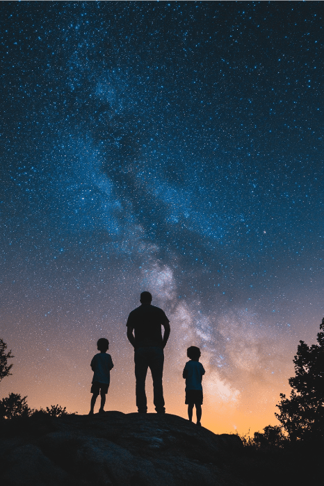 Las siluetas de un hombre y dos niños, de pie sobre una roca, contemplan el cielo nocturno estrellado con la Vía Láctea visible, una escena que recuerda a los poemas del Día del Padre para la iglesia, celebrando el vínculo bajo un vasto lienzo celeste.