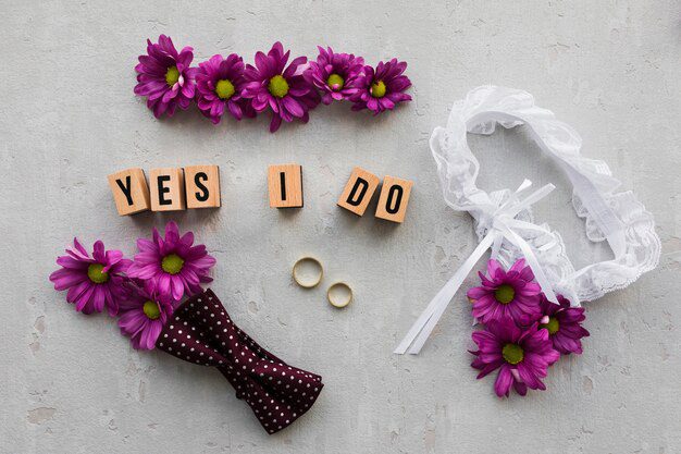 O tema do casamento apresenta flores roxas, azulejos "Yes I Do", uma liga de renda, duas alianças e uma gravata borboleta de bolinhas em uma superfície cinza. Para aqueles que estão pensando em "O que a Bíblia diz sobre o casamento após o divórcio?", essa pode ser uma conversa interessante para complementar o dia.