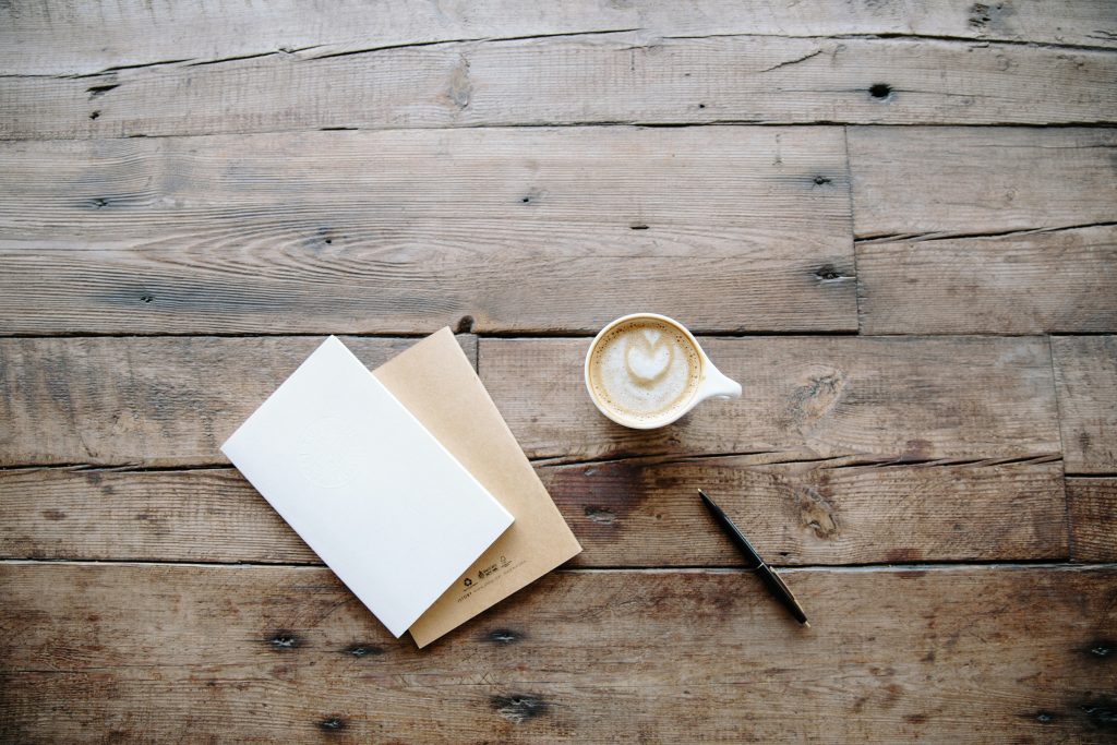Um caderno, uma caneta e um café com leite com a arte do coração repousam em uma mesa de madeira rústica, criando um ambiente sereno ideal para reflexão - talvez contemplando temas de consagração na Bíblia.