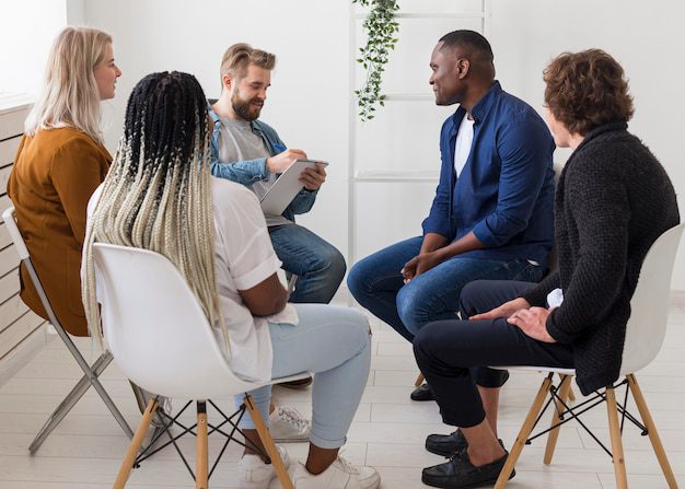Um grupo diversificado de pessoas sentadas em um círculo, engajadas em uma discussão sobre uma lição bíblica do Dia dos Pais em uma sala iluminada com decoração minimalista.
