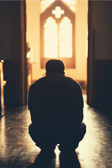 En una habitación iluminada por el sol, una persona con silueta se arrodilla, perdida en devociones para los líderes de la iglesia, mientras los vibrantes tonos de una vidriera proyectan coloridos reflejos a su alrededor.