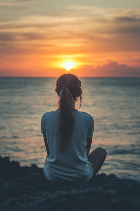 Una persona sentada en las rocas, contemplando una puesta de sol sobre el océano bajo un cielo nublado, como si encarnara una plegaria de AA contra la ansiedad: encontrar la paz en la serenidad y la inmensidad de la naturaleza.