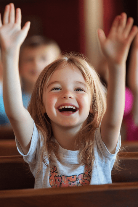 Un niño de pelo castaño levanta alegremente las manos mientras está sentado, sonriendo alegremente, irradiando la misma emoción que cuando se nombran nuevas clases para la iglesia infantil.