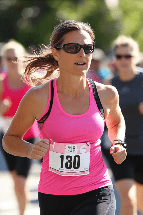 Una mujer, ejemplo de energía creativa, corre hacia delante con un top rosa vibrante y gafas de sol, llevando con orgullo el dorsal 180. Detrás de ella, otros corredores se suman al ambiente animado, cada uno de ellos parte de este dinámico evento juvenil. Detrás de ella, otros corredores se suman a la animada atmósfera, cada uno de ellos parte de este dinámico evento juvenil.