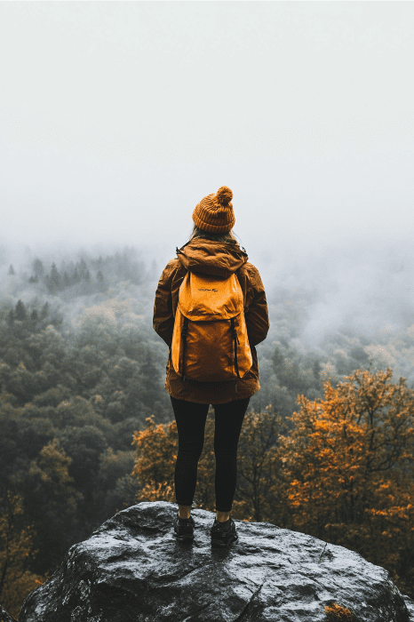 Una persona vestida con una chaqueta naranja y un gorro está de pie sobre una roca, con vistas a un bosque neblinoso con follaje otoñal, mientras reflexiona sobre ideas innovadoras para la captación de fieles.