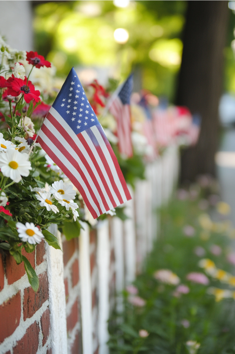 Pequeñas banderas americanas y flores adornan una pared de ladrillo, capturando la esencia del Día de la Independencia con un encanto sereno sobre un fondo borroso al aire libre. Perfecto para inspirar ideas de servicios religiosos que celebren la libertad y la unidad.
