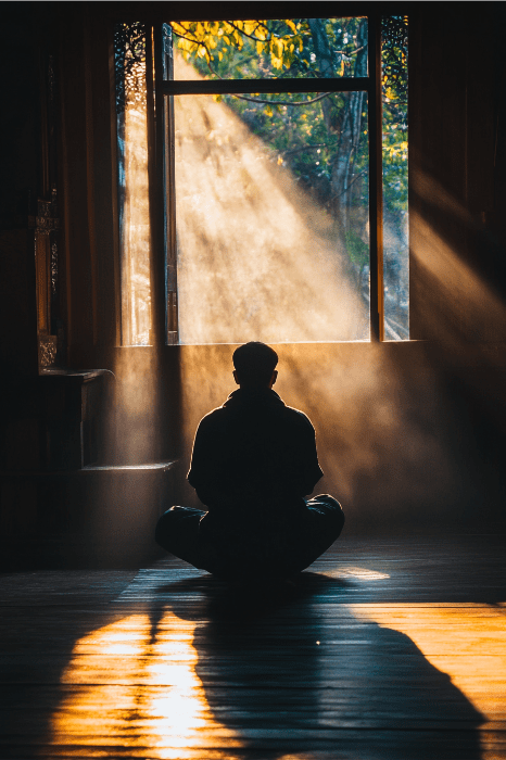 Silueta de una persona meditando en el interior, con la luz del sol entrando por una ventana y proyectando sombras dramáticas, como la reflexión serena que se encuentra en lo que es una sencilla oración diaria de AA, para encontrar la paz en medio de los desafíos de la vida.