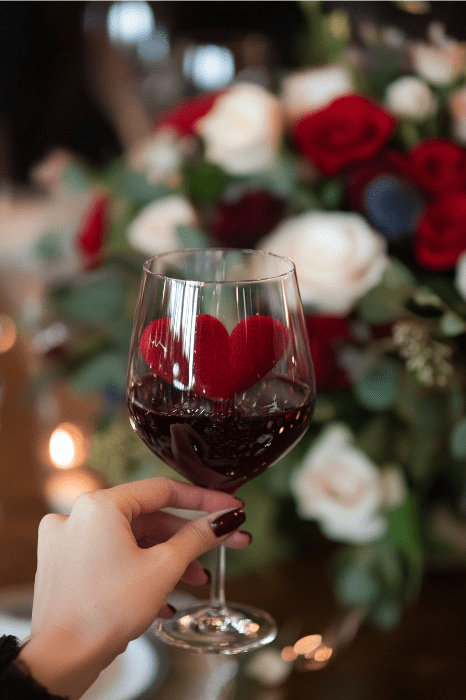 Uma mão segura uma taça de vinho tinto adornada com o desenho de um coração, enquanto rosas vermelhas e brancas se misturam suavemente ao fundo - um momento de tranquilidade, ecoando o calor da gratidão. Obrigado por visitar nossa igreja e compartilhar essa beleza serena.