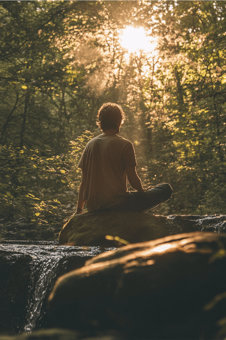 Una persona medita sobre una roca en un bosque iluminado por el sol, inmersa en una de las muchas actividades juveniles para el crecimiento espiritual, mientras la luz que fluye crea una atmósfera serena.