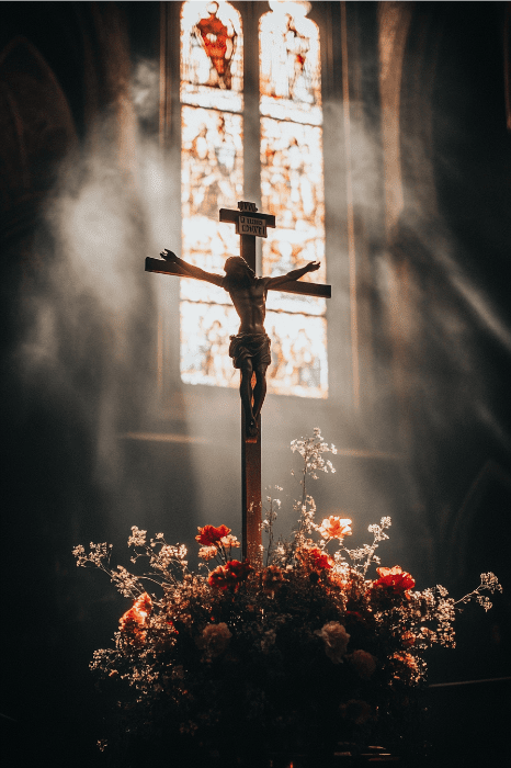 Crucifijo con flores debajo, bañado por la luz del sol a través de una vidriera, creando un entorno sereno para oraciones y reflexiones tranquilas.