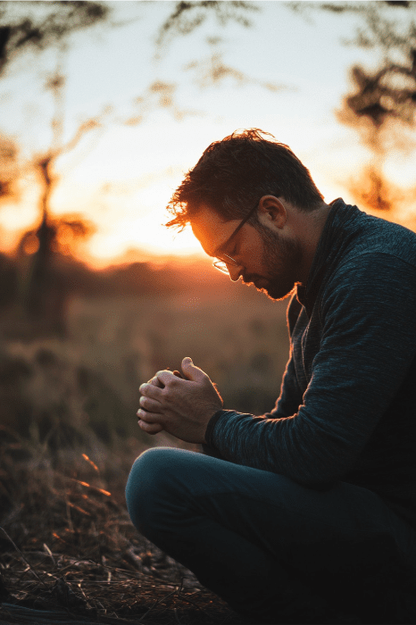 A man kneels in a field at sunset, hands clasped and head bowed, offering a quiet prayer for church growth.