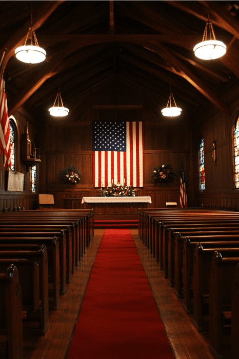 Vista interior de una iglesia, adornada con bancos de madera, una gran bandera de EE.UU. y vidrieras, iluminada por candelabros redondos: ideal para inspirar ideas para el servicio religioso del Día de la Independencia.
