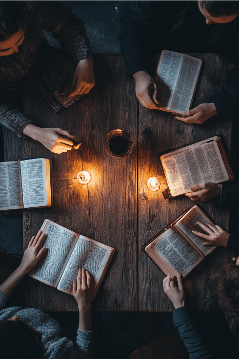 Cinco jóvenes creativos absortos en sus libros se reúnen en torno a una mesa de madera, rodeados de velas parpadeantes y una bebida en el centro, para compartir ideas de servicio e inspiración.