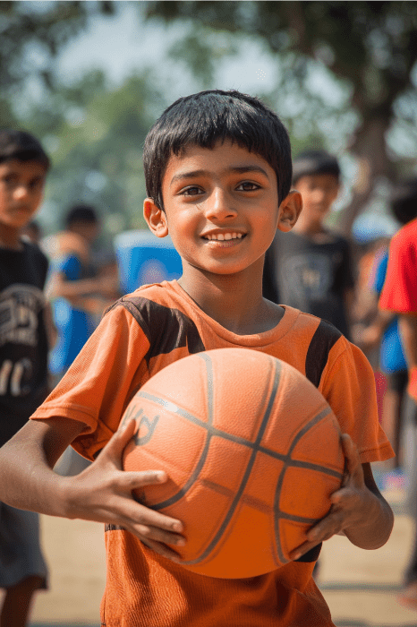 Un niño sonriente sostiene una pelota de baloncesto al aire libre, con otros niños jugando al fondo, lo que suscita ideas creativas entre los jóvenes para realizar actividades comunitarias atractivas.