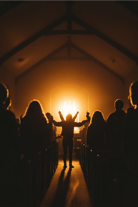 Una persona en silueta con los brazos levantados en una iglesia poco iluminada encarna la declaración de visión de las iglesias, de pie ante una luz brillante cerca del altar mientras otros se reúnen a su alrededor.