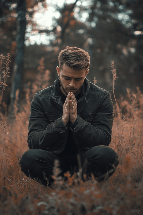 In a serene forest, a man kneels in prayer, hands clasped as if drawing strength from the scriptures for husbands and fathers. Clad in a dark jacket, he is encircled by vibrant autumn foliage, finding solace and guidance amidst nature's embrace.