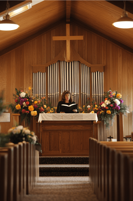 Uma mulher faz leituras para o Dia das Mães no púlpito de uma igreja, cercada por belos arranjos florais, com as notas suaves de um órgão tocando ao fundo.