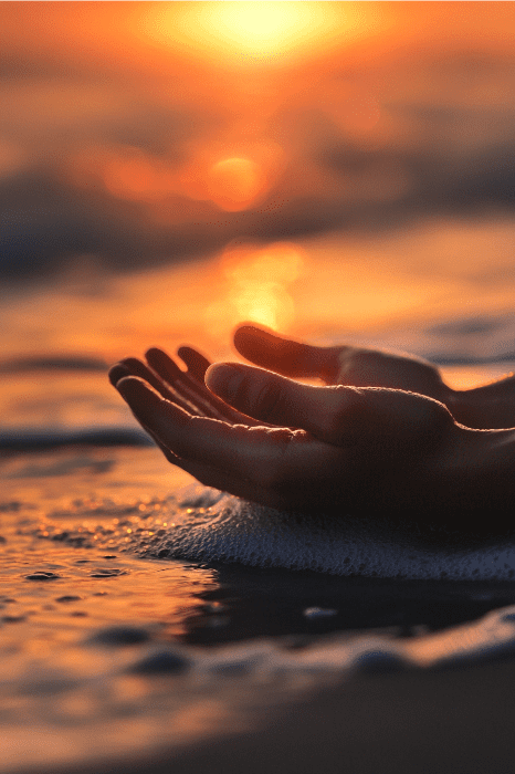 Manos ahuecando agua frente a una puesta de sol sobre el océano crean un momento de serenidad, que recuerda a las oraciones de Alcohólicos Anónimos, símbolo de limpieza y renovación.