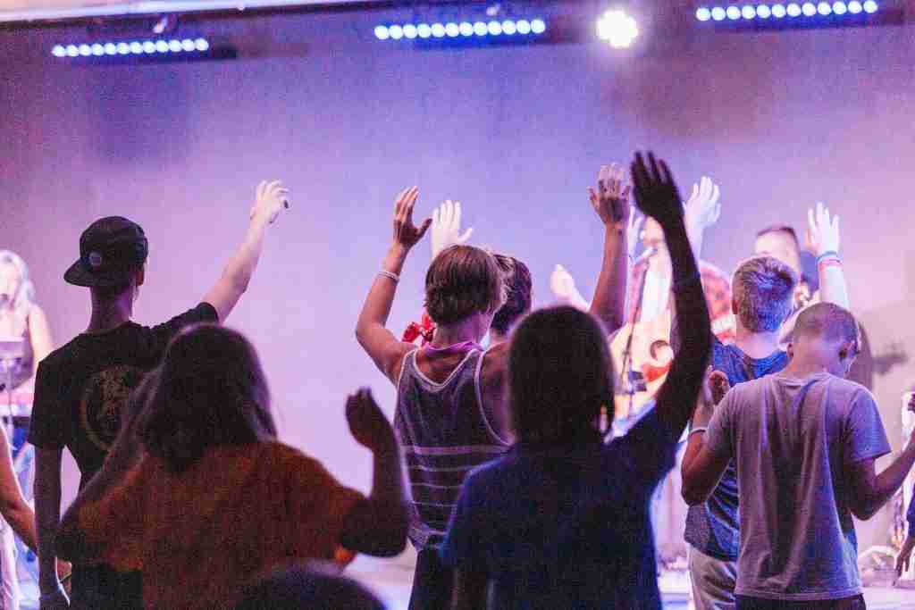 Bajo luces vibrantes y coloridas, una multitud se levanta con las manos en alto en un concierto, lo que recuerda el entusiasmo y la unidad que se sintieron durante una inspiradora lección de escuela dominical sobre la oración para los jóvenes.