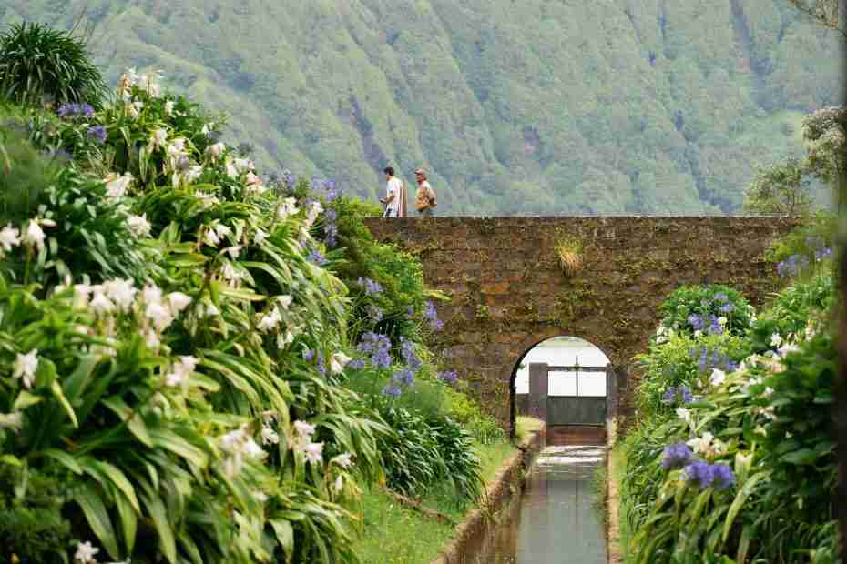 Dos personas caminan por un puente de piedra sobre un frondoso jardín, que recuerda a una manualidad de la escuela dominical Adán y Eva, con flores blancas y moradas, enmarcado por verdes montañas.