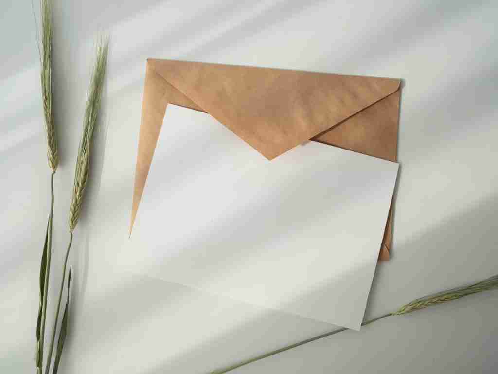 Una tarjeta blanca en blanco parcialmente dentro de un sobre marrón, con la inscripción sutil "AA prayer for anxiety", descansa junto a tallos de trigo sobre un fondo claro.