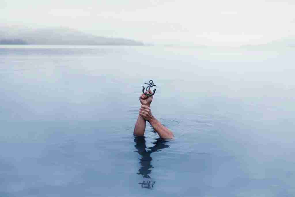 Los brazos de una persona se elevan desde aguas tranquilas sosteniendo una pequeña ancla metálica, símbolo de una oración de AA para la ansiedad, con montañas brumosas de fondo.
