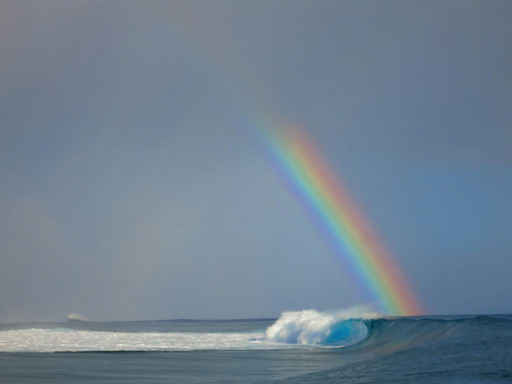 A rainbow, reminiscent of the symbolic rainbows in the Bible, arches over a blue ocean, with waves crashing under a cloudy sky.