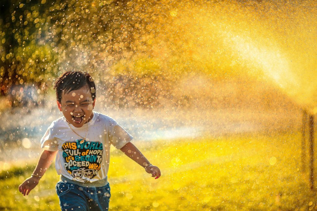 A todler boy runs through a sprinkler, showing the joy of thankfulness that a SUnday school lesson can teach