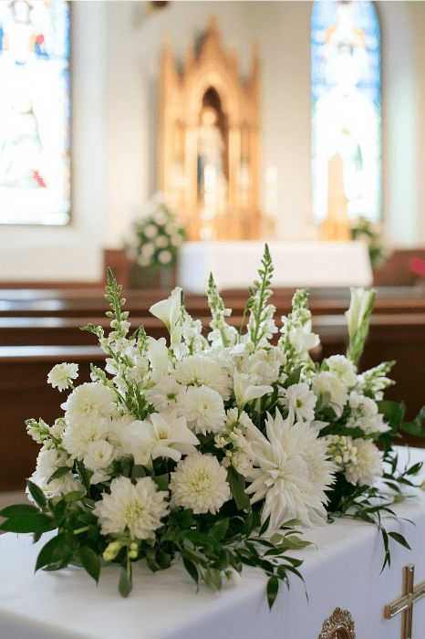 Explore impresionantes ideas de arreglos florales para altares de iglesias con este elegante despliegue floral en blanco, perfectamente colocado sobre el telón de fondo de unas vidrieras difuminadas.